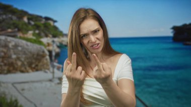 Kadın iki orta parmağını da parlak kumlu sahilde kayalık sahile doğru uzatıyor..