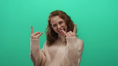 Kıvırcık saçlı bir kadın neşeli bir şekilde yeşil bir zemine taş el hareketi yapıyor, gözlüklü, sıradan örgü kazağıyla heyecan ve eğlenceyi ifade ediyor..