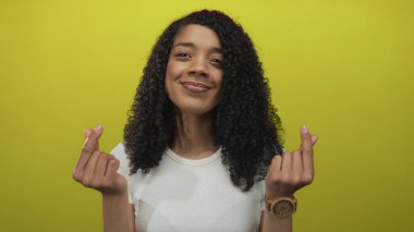 Beyaz tişört ve ahşap saat giyen Afrikalı Amerikalı kadın stüdyoda parmak kalp hareketi yapıyor ve geniş bir gülümseme sergiliyor..