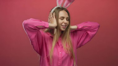 Tavşan kulaklı bir kadın, parlak pembe bir gömlek giyerken hayal kırıklığını ifade eden, canlı bir kırmızı arka plana karşı kulaklarını kapatıyor, neşeli tatil ruhunu ve mevsimsel neşesini sergiliyor.