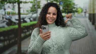 Genç İspanyol kadın kendini göstererek gülümsüyor ve açık bir sokak ortamında elinde bir akıllı telefon tutuyor, bulanık bir şehir arka planına karşı örülü bir süveter giyiyor..