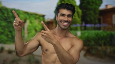 Tişörtsüz adam, yeşil yeşillik parkı dekoruyla kaplı binada geniş bir sırıtışla yukarı doğru işaret ediyor. Mutluluk..