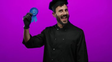 Siyah şef üniformalı İspanyol adam, mor arka plana karşı birinci sınıf mavi kurdeleyle kutlama yapıyor, neşe ve başarıyı ifade ediyor..