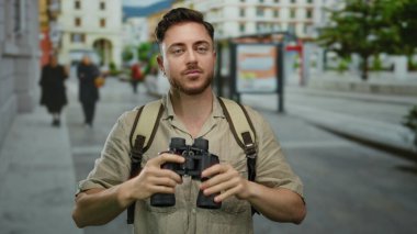 Şehrin hareketli sokaklarında dürbün kullanan sakallı genç bir adam, açık bir gökyüzünün altında sırt çantasıyla şehir ortamını keşfeden meraklı bir turisti gösteriyor..