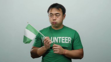 Beyaz arka planda Nijerya bayrağı taşıyan bir adam yeşil gönüllü tişörtü giyiyor ve ciddi bir ifade takınıyor..