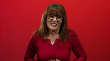 Kırmızı süveter giyen İspanyol kıdemli kadın canlı kırmızı bir duvar arkasında izole edilmiş. Rahatsız edici ama etkileyici görünüyor. Yüz ifadesi ve vücut dili ile duygularını etkili bir şekilde aktarıyor..