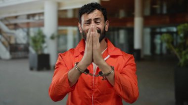 Turuncu tulumlu ve kelepçeli İspanyol bir adam bir otel lobisinde duruyor, açık havada yalvararak dua ediyor..
