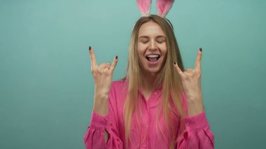 Paskalya tavşanı kulaklı genç bir kadın izole edilmiş yeşil bir duvarın arkasında neşe içinde poz veriyor. Mutluluğu ve eğlenceyi elleriyle ifade ediyor ve gülümsüyor..
