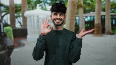 Sakallı genç adam gülümsüyor. Güneşli bir şehir caddesinde dikiliyor. Arka planda ağaçlar var..