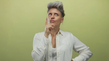 Kadın, profesyonel stüdyoda yumuşak yeşil zemin ile düşünceli pozda çeneye dokunur; merak içgözlem.