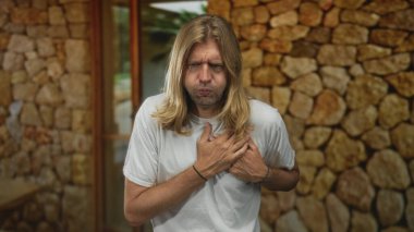 Uzun saçlı sarışın adam, taş duvarla çerçevelenmiş giriş kapısında dururken kalp krizi geçirmek için göğsünü sıkıyor..