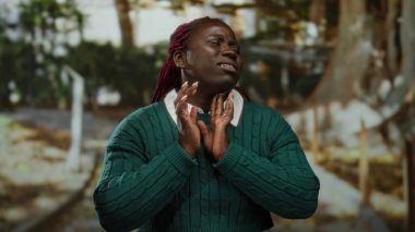 Yeşil kazaklı bir kadın parkta gergin bir ifadeyle boynuna dokunuyor. Bulanık ağaçlar ve patikalarla çevrili nefes almakta güçlük çekiyor..