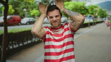 Dövmeli bir adam, şehir sokaklarında komik surat yapıyor güneşli şehir ortamında ağaçlar ve arka planda park edilmiş arabalarla çizgili tişört giyiyor..