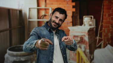 Sakallı İspanyol bir adam bir inşaat alanında güler yüzlü ve samimi bir şekilde işaret eder. Arka planda tuğlalı ve aletli bir kot ceket giyer. Sıradan ve ilgi çekici bir sahne yaratır..