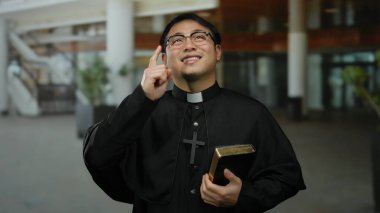 Siyah cüppeli Asyalı rahip elinde bir İncil tutuyor ve şehir ortamında gülümserken yukarı doğru işaret ediyor. İnanç ve maneviyata işaret ediyor..
