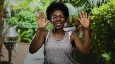 Açık hava parkında enerjik bir şekilde gülümseyen bir kadın, yemyeşil ile çevrili, neşeli ellerle sevinç ifade eden, neşeli, güneşli bir günde atmosfer yaratan bir kadın..