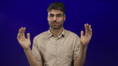 Latin kökenli genç adam gözlük takıyor ve avuçlarını mavi bir stüdyoda bej bir gömlek giyerek ayırıyor..
