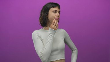 Açık gri gömlekli genç İspanyol kadın mor stüdyoda hafif kaşları ve çilleri olan çeneye dokunur..
