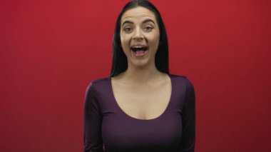 Kadın, elleri açık bir şekilde kırmızı duvara karşı gizemli bir jest yapıyor. Neşeli bir ifade ve renkli bir ortam sergiliyor..