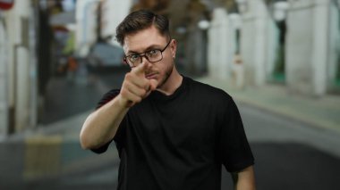 Sakallı ve gözlüklü genç bir adam şehir merkezinde siyah gömlek giyip, tasvip etmediğini belirterek, bir şehir caddesinde küçümseyici bir jest yapıyor..