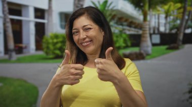 Sarı bluz giyen İspanyol orta yaşlı kadın, şehir sokağında palmiye ağaçları ve bina cephesiyle dolu sıcak bir gülümsemeyle çifte onay veriyor; onay pozitifliği iyimserliği teşvik ediyor.