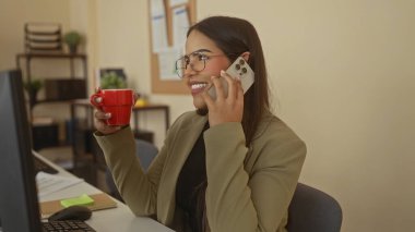 Elinde kırmızı bardakla gülümseyen ve bir ofis ortamında telefonla konuşan, görünür rafları ve masasında bir bilgisayarla verimli bir çalışma ortamı sunan bir kadın..