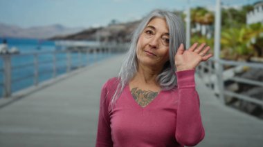 Gri saçlı, pembe gömlekli, deniz kenarında gezinen açık gökyüzü altında kumsal ve okyanus manzaralı bir kadın..