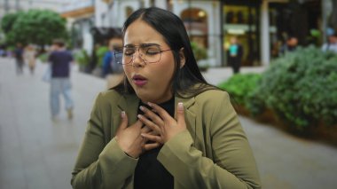 Meşgul bir caddede göğsünü sıkıca tutarak öksüren bir kadın. Genç Latin asıllı, açık havada acı çeken bir kadın ise acılı bir tavır takınıyor..