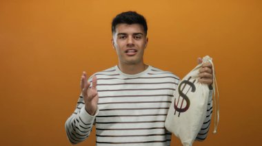 Turuncu arkaplan önünde para çantası tutan genç adam İspanyol kökenli ve sıradan çizgili giyim tarzını sergiliyor, finansal başarısını ifade ediyor..