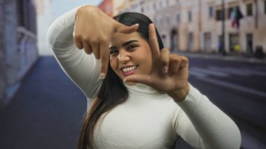 Açık havada gülümseyen beyaz kazak giyen, uzun saçlı, elleriyle yüzünü çerçeveleyen bir kadın şehir sokaklarının arka planında mutluluk ve yaratıcılığı taşıyor.