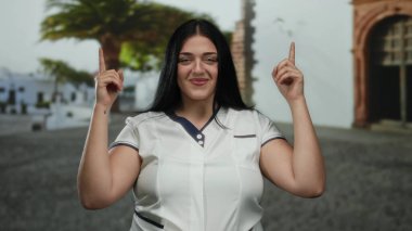 Açık havada gülümseyen bir kadın işaret parmaklarıyla şehrin arka planına doğru bakıyor..