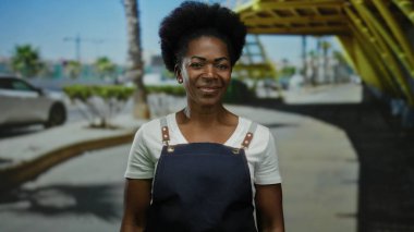 Şehir caddesinde gülümseyen bir kadın modern şehir arkaplanı ve canlı renklerle önlük giyerek açık hava ortamının çeşitliliğini ve enerjisini vurguluyor..