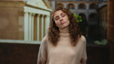 Eski bir üniversitede gözlüklü ve kıvırcık saçlı bir süveter giyen bir kadın klasik mimariyle çevrili düşünceli bir ifade sergiliyor..