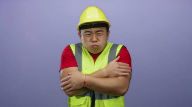 Kask ve güvenlik yeleği giyen genç adam, izole edilmiş mor bir arka plan önünde soğuk görünüyor. Rahatsızlık ve ürperti hislerini iletiyor..