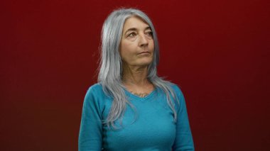 Gri saçlı yaşlı bir kadın canlı kırmızı bir arka plana karşı güvenle duruyor, bilgeliğini ve zarafetini basit turkuaz kıyafetleriyle sergiliyor, izole ve odaklanmış..