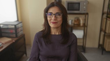 Gözlüklü kadın, ofis binasının arkasında istiflenmiş depolama kutuları ve mikrodalga fırınla masada oturuyor..