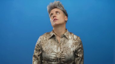 Kısa gri saçlı, yılan desenli bluz giyen beyaz kadın mavi stüdyoda gözleri yuvarlıyor..