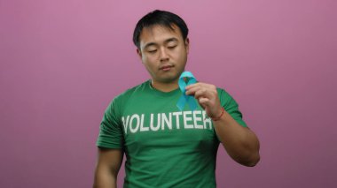 Yeşil tişörtlü genç Çinli gönüllü, kanser farkındalığını ve desteğini simgeleyen pembe arka planda mavi kurdele tutuyor..