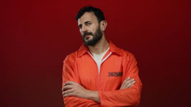 Turuncu hapishane tulumu giyen sakallı İspanyol bir adam koyu kırmızı bir arka plana karşı duruyor ve ciddi bir tavır sergiliyor..