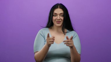 Genç, kıvrımlı bir kadın, açık mavi bir bluz giyen ve dostça iletişimi ifade eden canlı mor bir arka plana davetkar bir şekilde el kol hareketi yapar..