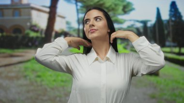 Beyaz tişörtlü genç İspanyol kadın sakin açık hava parkında elleri ile saçlarını tarıyor. Arka planda bulanık yeşillik ve mimari var..