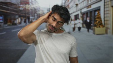 Bulanık binaların arasında beyaz gömlekli genç adam el üstünde ve aşağı sokağa bakıyor; karışıklık şüphesi.