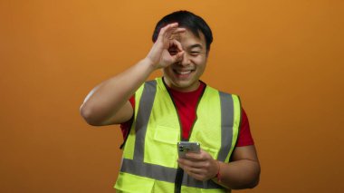 Adam gülümsüyor ve turuncu arka planda yüksek görünürlüklü yelek giyiyor.