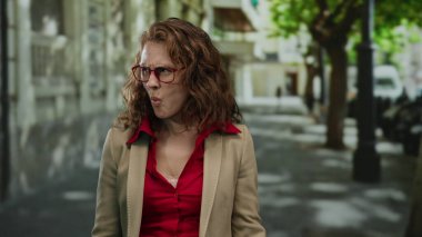 Kırmızı gözlüklü ve bej ceketli bir kadın, güneşli bir günde şehrin arka planına bakarak şaşkınlık ve şaşkınlık gösteren bir şehir caddesinde duruyor..