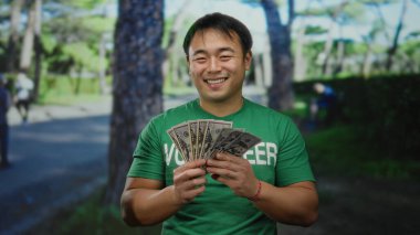 Sokak ortasında elinde Amerikan doları tutan genç bir adam yeşil bir gönüllü tişört giyerken açık hava şehrinde çekici bir şekilde gülümsüyor..