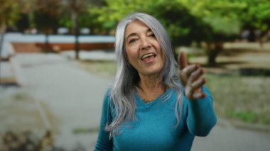 Gri saçlı yaşlı kadın elini uzatıyor ve yeşil ağaçlar ve yürüyüş yolu olan bir parkta neşeyle gülümsüyor..