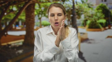 Yeşil bir parkta üşüyen bir kadın beyaz bir gömlek giyiyor, sarı saçlı, açık havada, doğada bir ürperti anı yakalıyor..