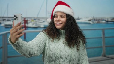 Noel Baba şapkalı bir kadın tatil sezonunda teknelerle deniz kenarında selfie çekiyor. Canlı bir açık hava manzarası ve şenlikli bir ruh hali sergiliyor..