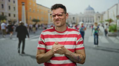 Vatikan şehir meydanında gözlüklü genç adam kırmızı çizgili tişört giyiyor. İnsanlar ve şehir geçmişi güneşli bir günde sokak modası ve modern dövme sergiliyor..