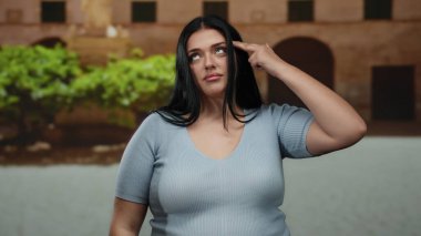 Şehir sokaklarında elini başını gösteren parmaklarıyla el kol hareketi yapan, hayal kırıklığını ya da tefekküre dalmayı ifade eden, artı boy ve kıvrımlı vücut, günlük mavi bluzlar giyen bir kadın..